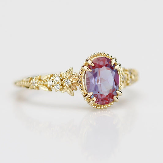 Vintage-inspired floral engagement ring in solid yellow gold featuring a color-changing lab alexandrite center stone. The ornate band is sculpted with gold leaves and flowers, accented by small white diamonds, showcasing a teal-purple gemstone on a white reflective surface.