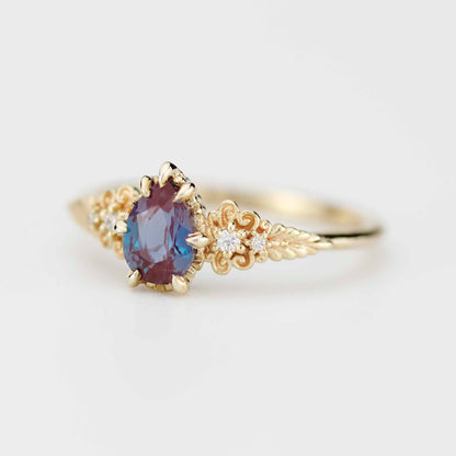 Elegant Nooi Jewelry gold ring featuring an oval-cut bi-color gemstone in red and blue tones, set in prongs with intricate filigree detailing and small accent diamonds along the band.