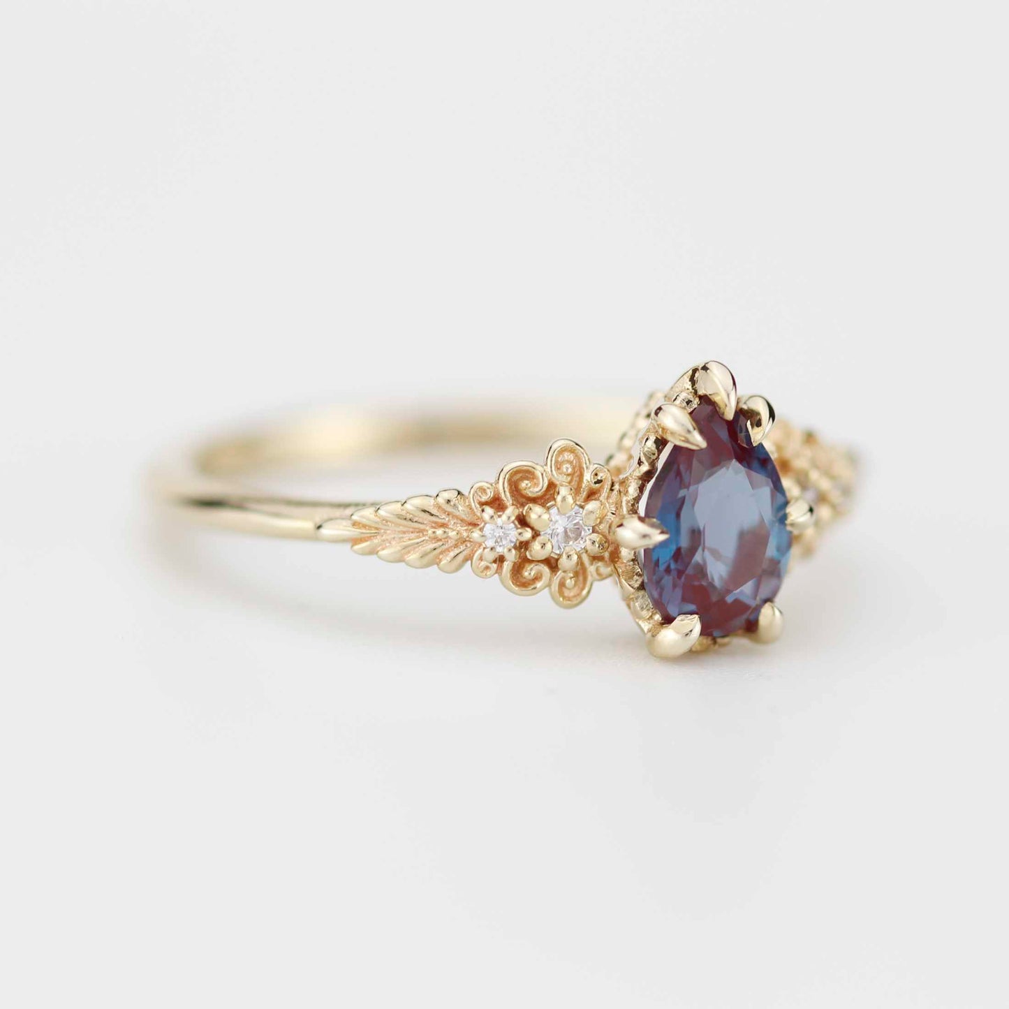 Elegant gold ring with a pear-shaped, color-shifting gemstone in deep blue and burgundy tones, set in intricate floral-inspired detailing with small diamond accents along the band.