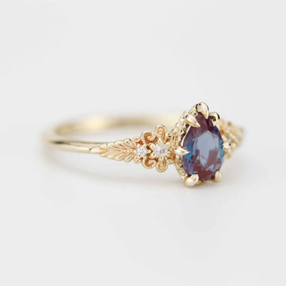 Elegant gold ring with a pear-shaped, color-shifting gemstone in deep blue and burgundy tones, set in intricate floral-inspired detailing with small diamond accents along the band.