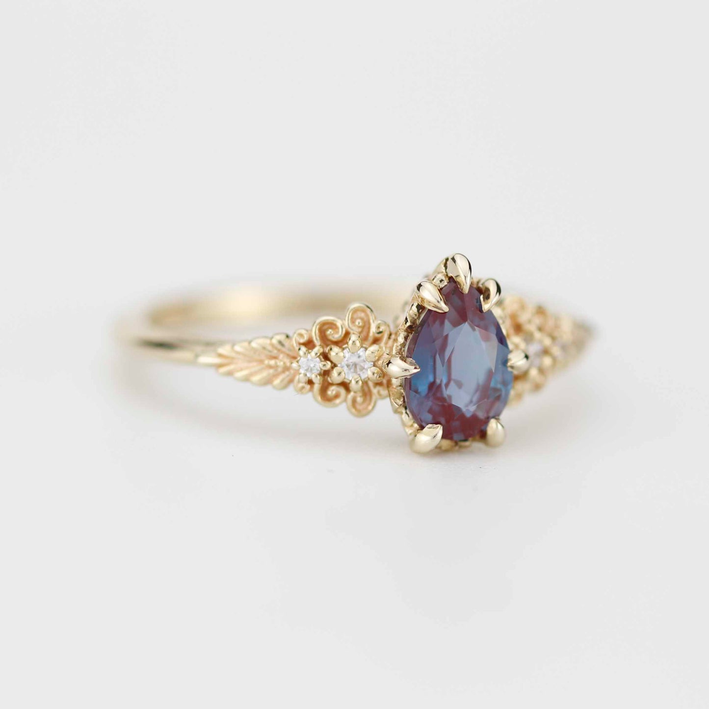 Delicate gold ring featuring a pear-shaped, multicolored gemstone that shifts between deep blue and reddish hues, set in ornate vintage-inspired detailing with small accent diamonds along the band.