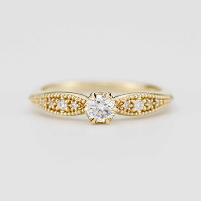 Close-up of a vintage-style 18K solid yellow gold ring featuring a round brilliant-cut natural diamond in a 6-prong setting. The tapered band is decorated with intricate milgrain beading and fine granulation textures, set against a soft beige background.