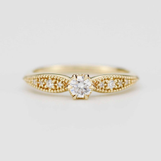Close-up of a vintage-style 18K solid yellow gold ring featuring a round brilliant-cut natural diamond in a 6-prong setting. The tapered band is decorated with intricate milgrain beading and fine granulation textures, set against a soft beige background.
