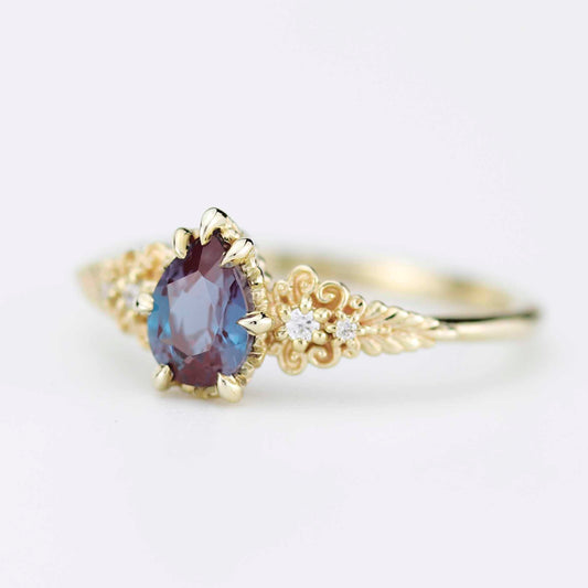 Detailed view of the Twilight Fern ring in 18k yellow gold, showcasing a pear-cut purple alexandrite stone held by claw prongs on a decorative filigree band with small diamond accents.
 
