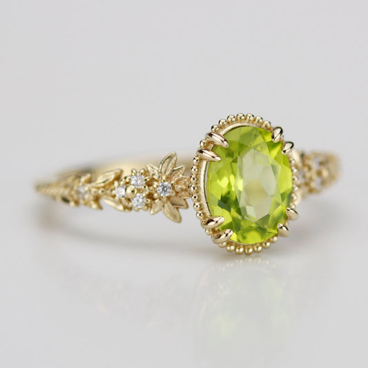 Oval-cut natural green peridot ring in 18k yellow gold with diamond-studded floral band details on a white reflective surface.