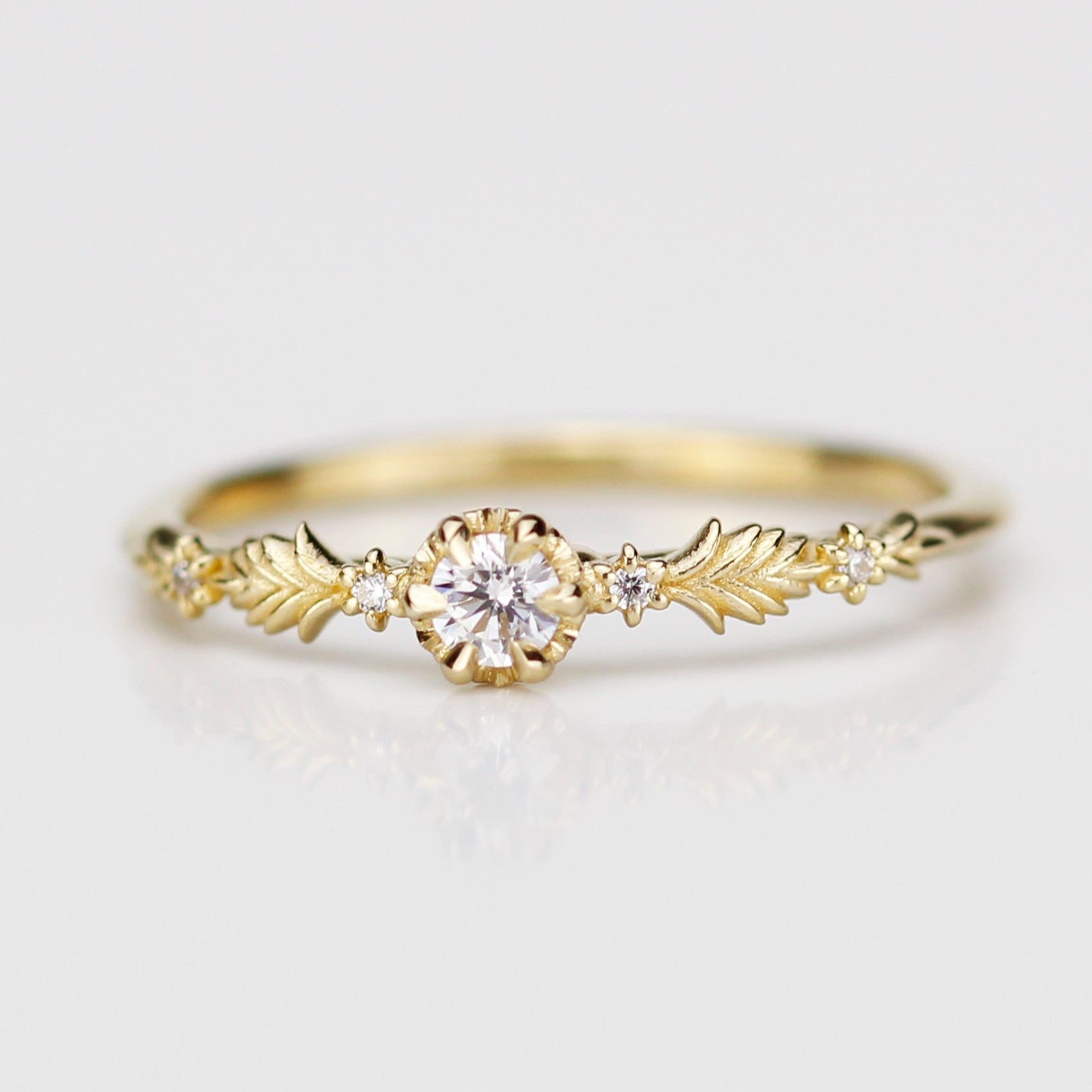 18k yellow gold Florisia ring featuring a central 3mm natural white diamond in a textured setting flanked by intricate gold leaf patterns and four tiny accent diamonds on a clean white reflective surface.