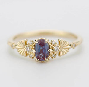 An elegant 18K gold ring featuring a 6x4 mm oval lab-created Alexandrite gemstone at the center, flanked by natural diamond accents and intricate floral-inspired detailing on the band, showcasing a vintage-inspired design.