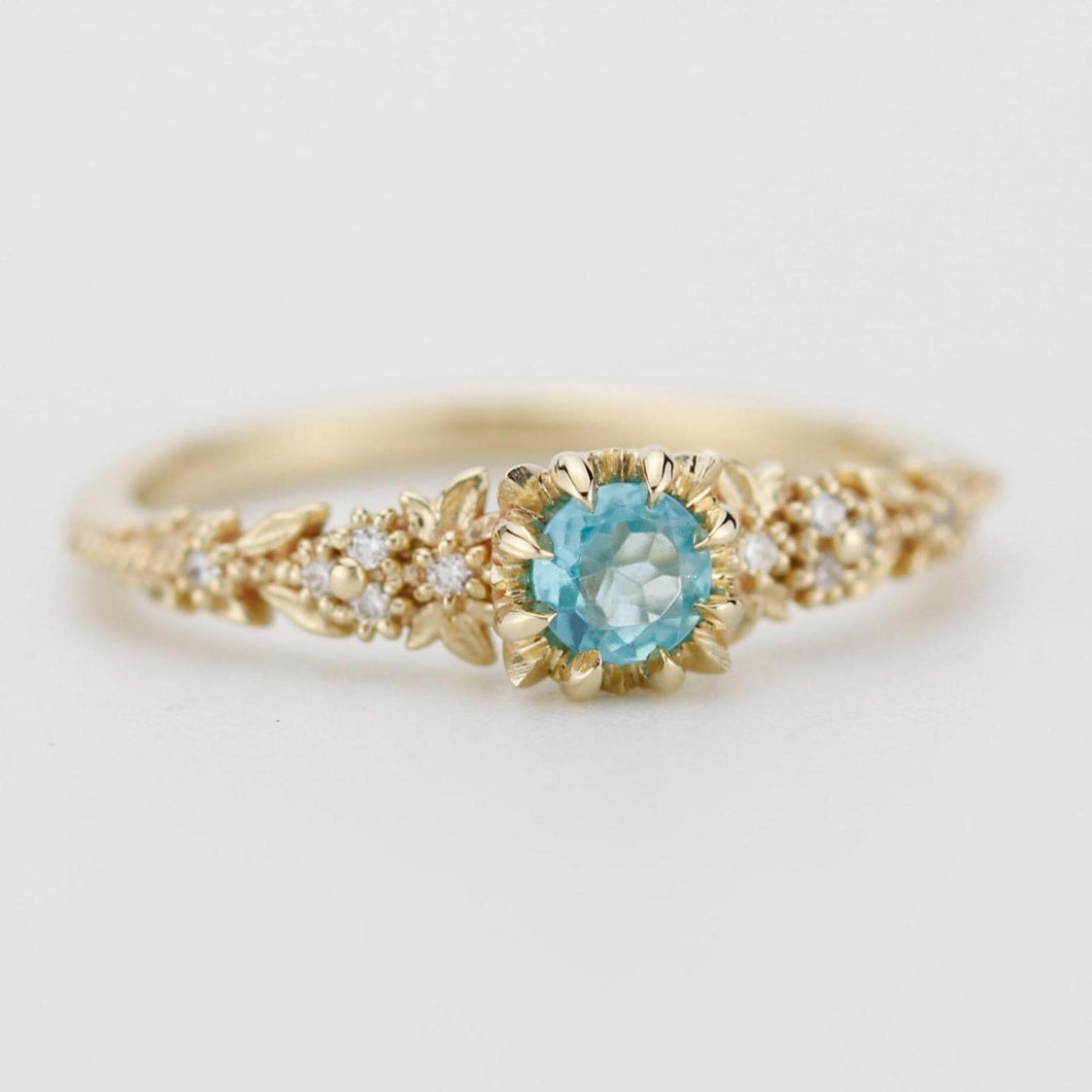 Delicate 18k gold engagement ring with a round blue apatite center stone and diamond accents in a floral setting.