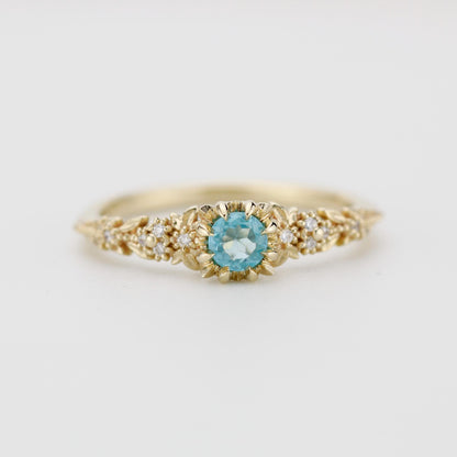 Delicate 18k gold engagement ring with a round blue apatite center stone and diamond accents in a floral setting.
