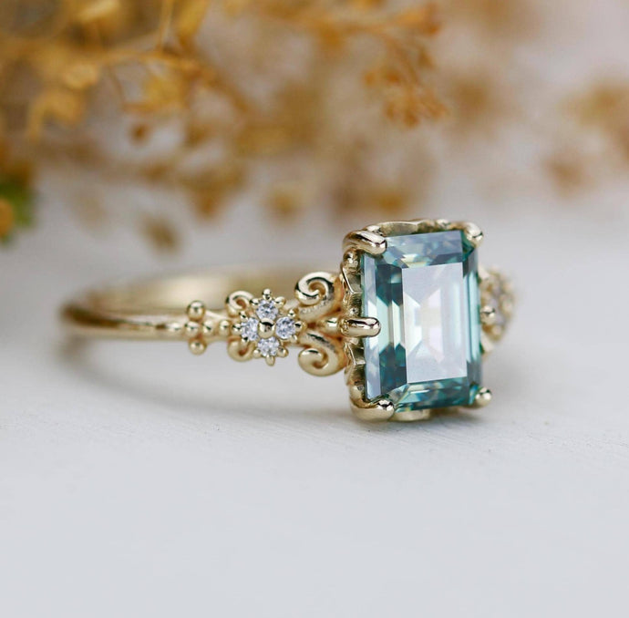 Vintage-style 18K gold ring featuring an emerald cut teal green moissanite with natural diamond accents and detailed filigree design.