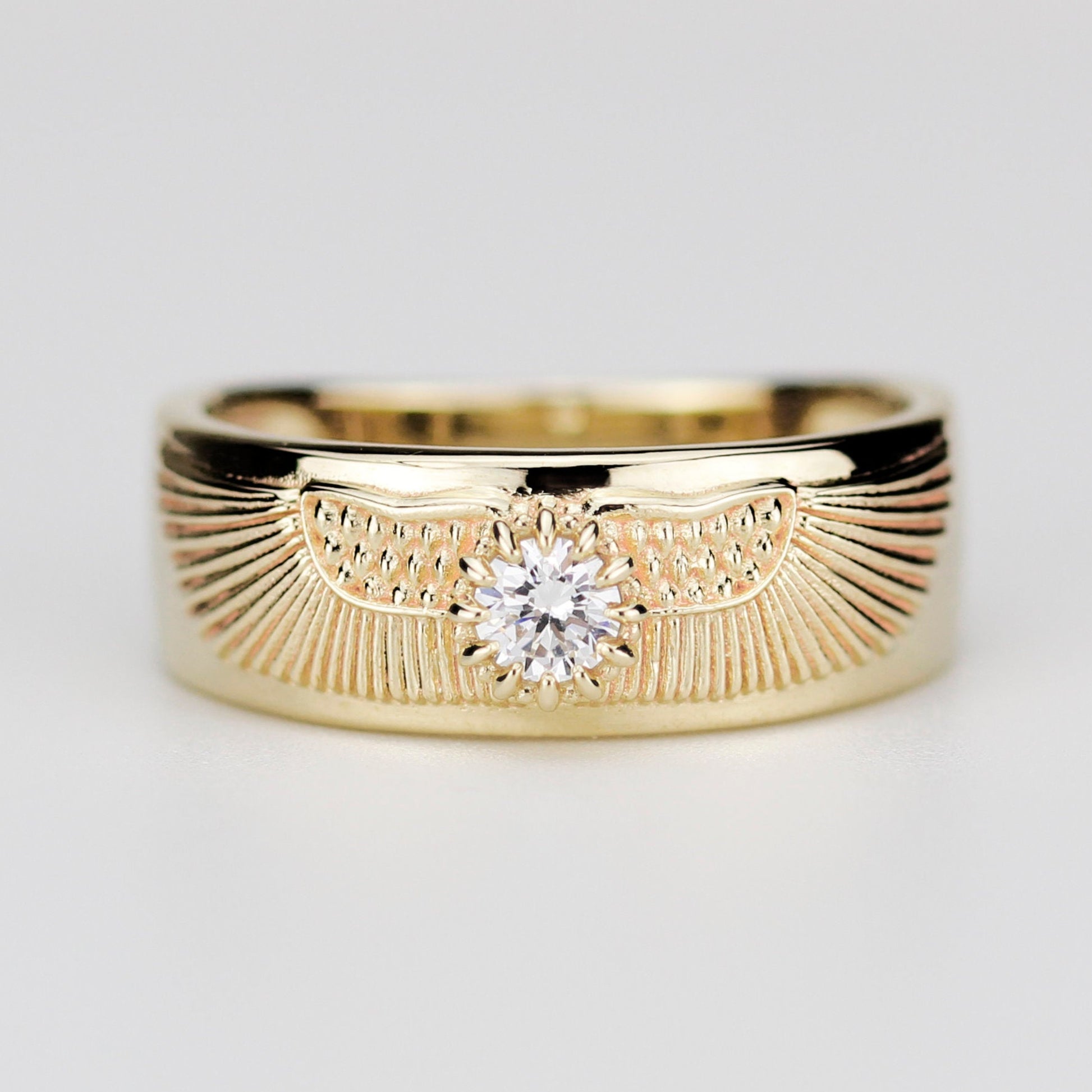 Centered close-up of a wide, solid 18K yellow gold band ring featuring a brilliant-cut natural diamond in the middle, flanked by detailed ancient Egyptian winged sun disk engravings on a plain grey background.