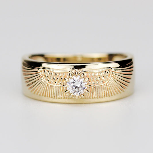 Centered close-up of a wide, solid 18K yellow gold band ring featuring a brilliant-cut natural diamond in the middle, flanked by detailed ancient Egyptian winged sun disk engravings on a plain grey background.
