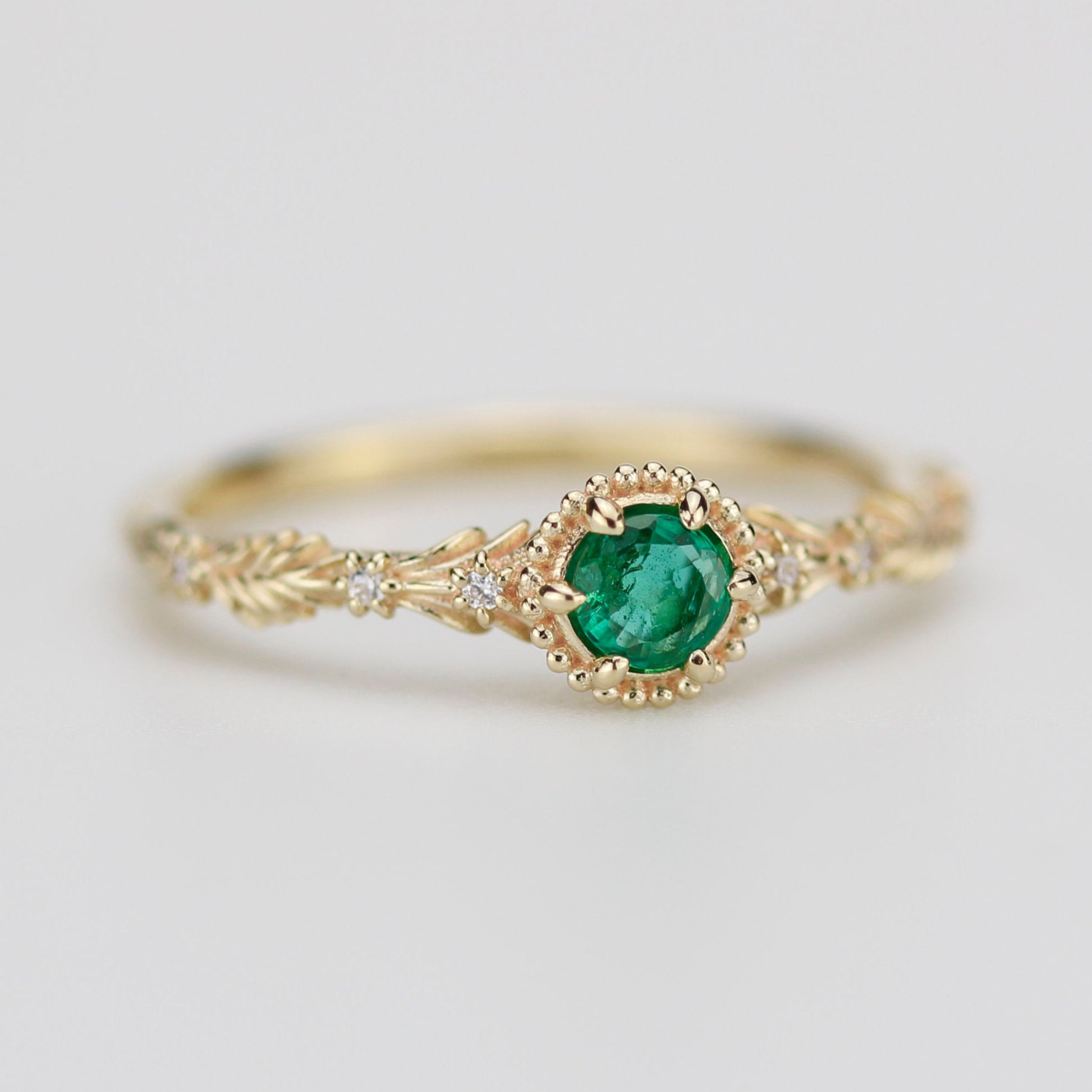 Close-up of an 18k gold botanical ring featuring a round green natural emerald center stone in a beaded bezel setting, with detailed leaf carvings on the band and three tiny diamonds on each side.