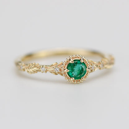 Close-up of an 18k gold botanical ring featuring a round green natural emerald center stone in a beaded bezel setting, with detailed leaf carvings on the band and three tiny diamonds on each side.